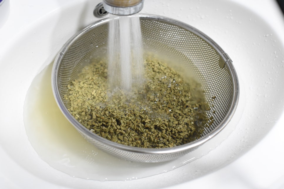 Hands rinsing filter media in a clear bowl under running water in a clean sink, lab-like setting, re